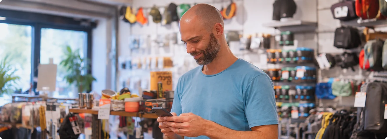 גבר קירח עם זקן ולבוש בחולצה כחולה מסתכל ומחייך אל מכשיר הטלפון הנייד שבידיו בתוך חנות.