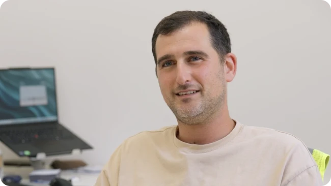 תצלום דיוקן של גבר צעיר המחייך למצלמה בסביבת משרד, כשברקע מונח מחשב נייד.